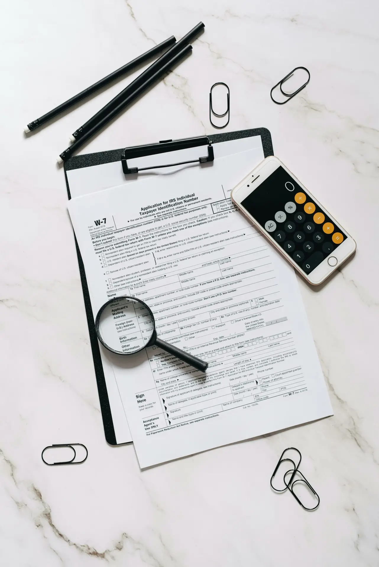 High angle shot of tax form, magnifying glass, and smartphone calculator on marble surface.